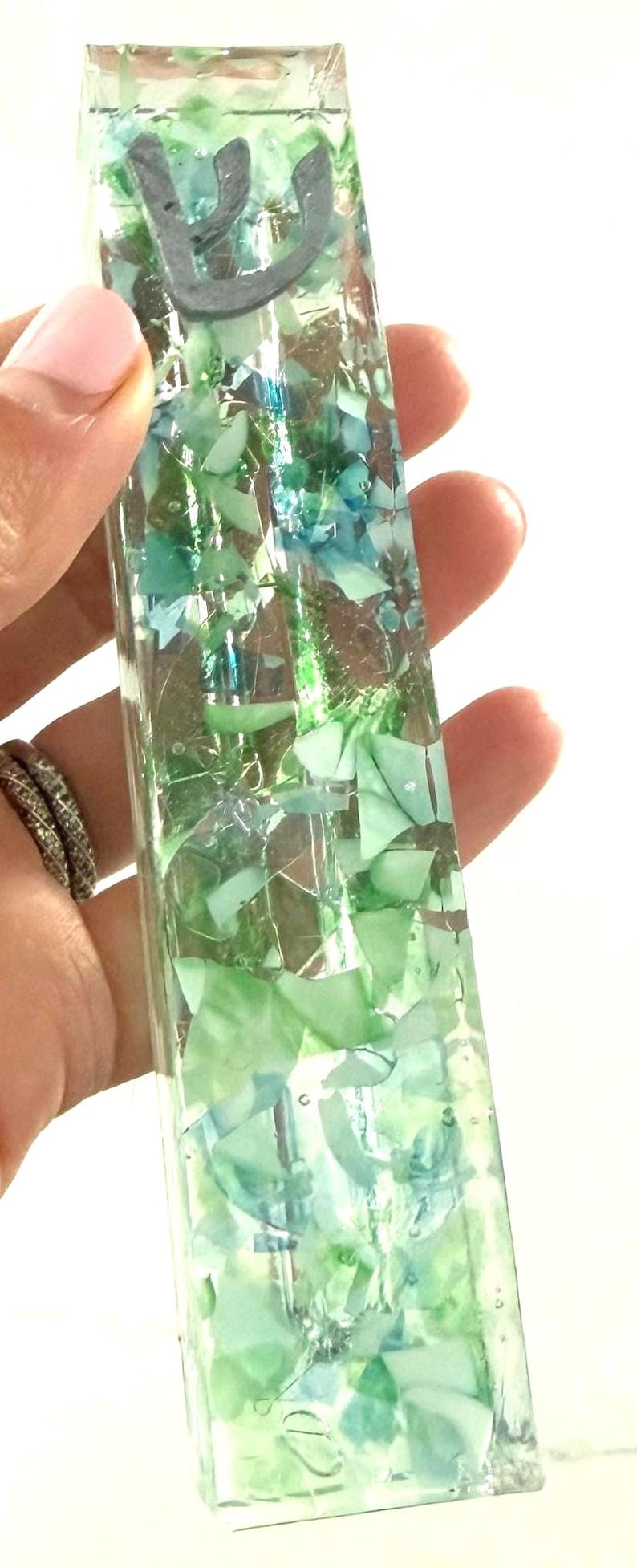 May include: A rectangular mezuzah case made of clear resin embedded with various shades of green and blue glass fragments. The top section features a dark gray Hebrew letter Shin, which is a traditional symbol found on this type of religious object.