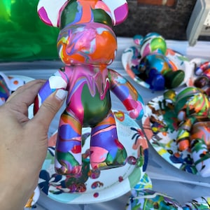 May include: A bear-shaped figurine featuring a glossy, multi-colored paint drip design. The surface displays a vibrant swirl of orange, pink, green, blue, and white colors in a fluid, marbled pattern across the head, torso, and limbs.