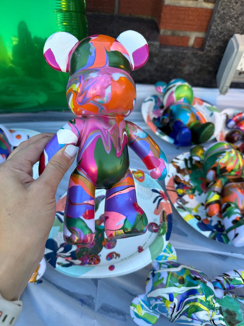 May include: A bear-shaped figurine featuring a glossy, multi-colored paint drip design. The surface displays a vibrant swirl of orange, pink, green, blue, and white colors in a fluid, marbled pattern across the head, torso, and limbs.