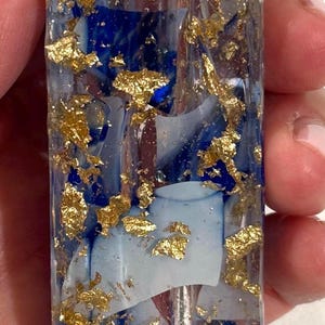 May include: A rectangular clear resin mezuzah case featuring embedded blue pigment swirls and scattered gold leaf flakes. A raised, textured gold-colored Hebrew letter Shin is positioned near the top of the transparent, elongated vertical structure.
