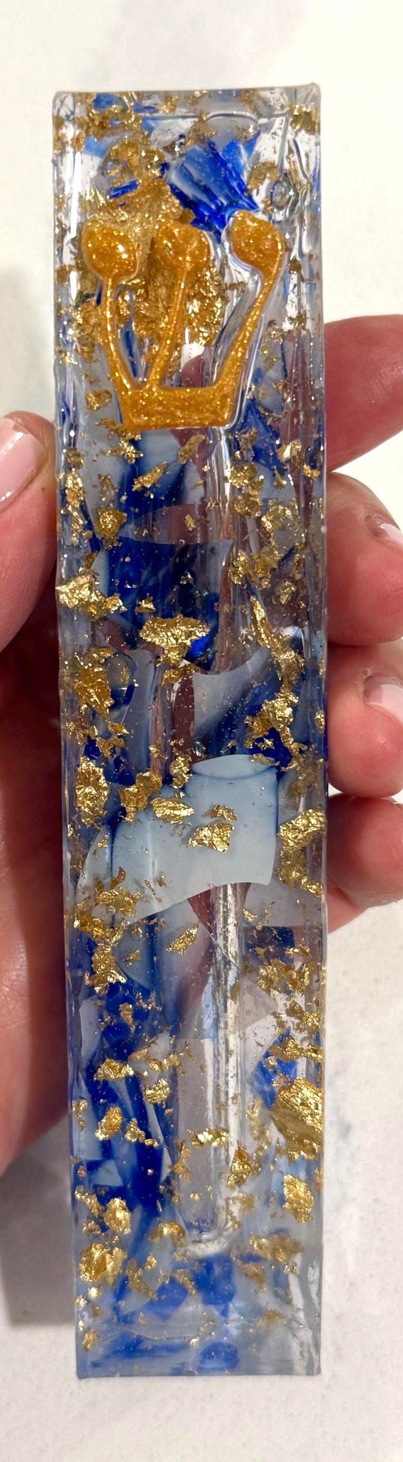May include: A rectangular clear resin mezuzah case featuring embedded blue pigment swirls and scattered gold leaf flakes. A raised, textured gold-colored Hebrew letter Shin is positioned near the top of the transparent, elongated vertical structure.