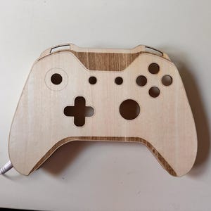 Könnte beinhalten: Ein holzfarbenes Objekt in Form eines Xbox-Controllers mit hellbraunem Finish. Der Controller hat ausgeschnittene Löcher für Tasten und Joysticks sowie ein kreuzförmiges Steuerkreuz. Ein weißes Kabel mit Stecker ragt seitlich heraus.