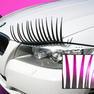 May include: A white car with black eyelashes applied to the headlights. The car has a black grill and a black bumper. The eyelashes are made of a black material and are designed to look like real eyelashes. The car is parked on a pink background.