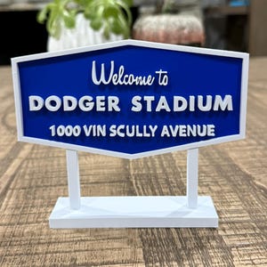 以下が含まれることがあります： 「Welcome to DODGER STADIUM 1000 VIN SCULLY AVENUE」と書かれた青と白のサイン。 白い縁取りのある六角形で、白いベースに2本の白い柱で支えられています。 装飾品です。