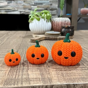 May include: Three orange, knitted pumpkins with black eyes and smiling mouths. Each pumpkin has a green stem. The pumpkins are different sizes and are displayed on a wooden table. The pumpkins are a fall or Halloween decoration.