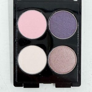 Puede incluir: Una paleta de sombras de ojos cuadrada de color negro que contiene cuatro recipientes circulares de polvo compacto. El recipiente superior izquierdo es rosa claro, el superior derecho es morado intenso, el inferior izquierdo es blanco roto y el inferior derecho es un tono marrón topo brillante.