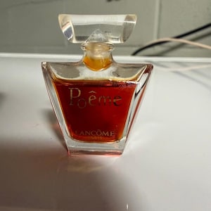 Puede incluir: Un frasco de perfume de vidrio transparente con una forma ancha y angular y un tapón transparente. El frasco contiene un líquido de color ámbar intenso y presenta el texto dorado Poême y Lancôme Paris impreso en la parte frontal del vidrio.