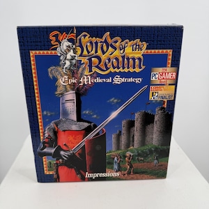 May include: A rectangular video game box for Lords of the Realm featuring a knight in red and black armour holding a sword. The cover displays a stone castle, a blue sky, and text including Epic Medieval Strategy and Impressions branding.