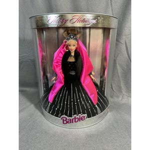 Puede incluir: Una muñeca Barbie en una caja cilíndrica transparente. La muñeca lleva un vestido negro con detalles plateados y un chal rosa. La caja lleva la etiqueta "Happy Holidays" y "Barbie".