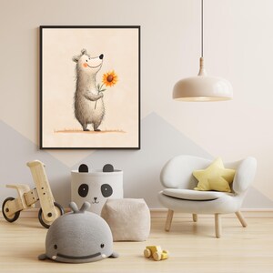 May include: A framed watercolor print of a smiling bear holding a sunflower. The artwork features soft colors and is displayed in a child's room with a wooden toy, a panda-themed storage bin, and a white chair with a yellow star pillow.