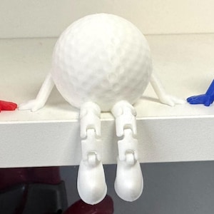Op de afbeelding: Drie golfbalfiguren in rood, wit en blauw, elk met armen en beweeglijke benen, zittend op een witte plank. De golfballen hebben een putjesstructuur en de benen zitten in een zittende positie.