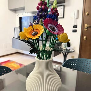 May include: A white, ribbed vase filled with colorful artificial flowers. The arrangement includes yellow, pink, and purple blooms, with green stems. The vase sits on a glass table, creating a vibrant home decor display.