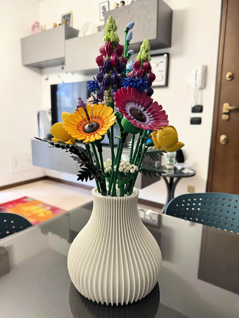May include: A white, ribbed vase filled with colorful artificial flowers. The arrangement includes yellow, pink, and purple blooms, with green stems. The vase sits on a glass table, creating a vibrant home decor display.