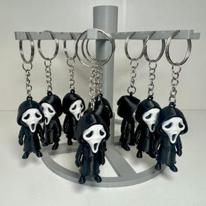 May include: A collection of black and white Ghostface keychains hanging on a gray display stand. Each keychain features a small, 3D-printed figure of the iconic Scream movie character, with a white mask and black robe.