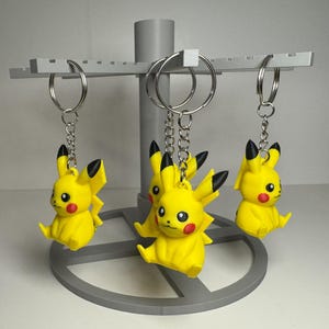 May include: Three yellow Pikachu keychains with black accents and red cheeks hang from a gray display stand. Each keychain has a silver ring and chain. The stand is gray and has a circular base.