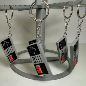 Peut inclure: Porte-clés gris avec un design de manette de jeu rétro. Les manettes ont des boutons noirs et des accents rouges, suspendues à des anneaux et chaînes argentés. Les porte-clés sont exposés sur un présentoir gris à plusieurs niveaux.