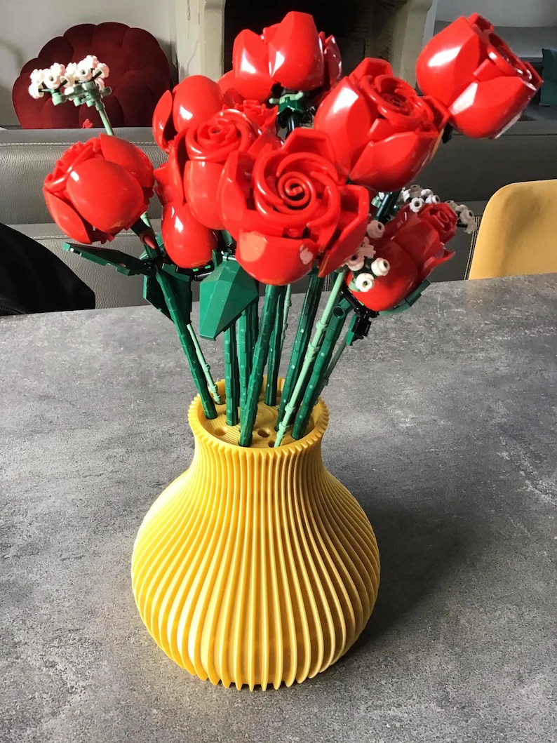 May include: A vibrant bouquet of red roses in a yellow vase. The roses are made of plastic and have green stems. The vase is a bright yellow color and has a ribbed design.