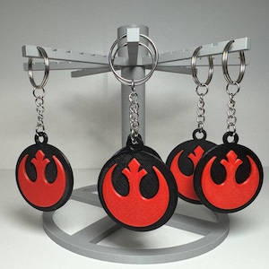 May include: A display stand holds five keychains. Each keychain has a black, circular pendant with a red symbol. The pendants are attached to silver chains and keyrings. The stand is gray and has a circular base.