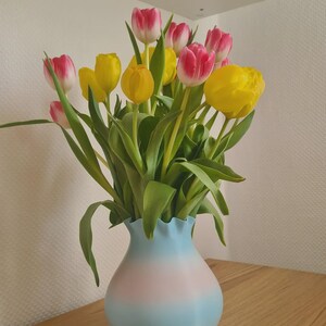 May include: A bouquet of tulips in a vase. The tulips have yellow and pink petals with green stems and leaves. The vase is light blue and pink, with a ruffled top. The arrangement sits on a wooden surface.