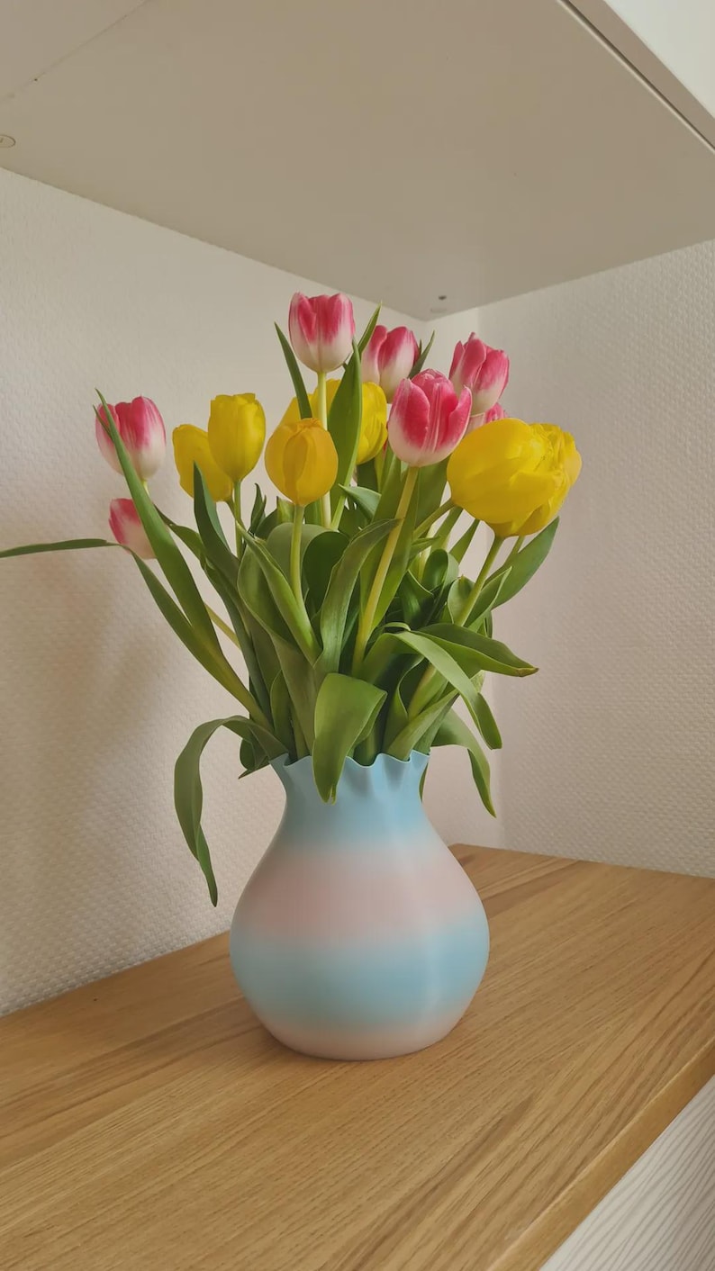 May include: A bouquet of tulips in a vase. The tulips have yellow and pink petals with green stems and leaves. The vase is light blue and pink, with a ruffled top. The arrangement sits on a wooden surface.