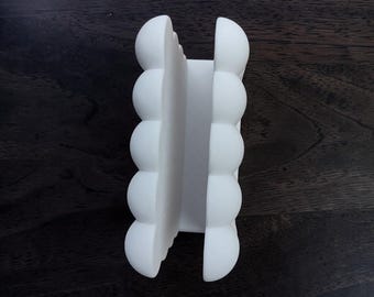 Puede incluir: Un servilletero de color blanco mate con un diseño vertical, compuesto por dos lados paralelos formados cada uno por cinco segmentos redondeados y semiesféricos apilados. El objeto tiene un acabado liso y un espacio central abierto.