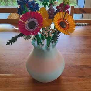 May include: A colorful bouquet of artificial flowers in a light blue and pink vase. The flowers include pink, yellow, and orange sunflowers, blue and purple blooms, and green stems. The vase sits on a wooden table.