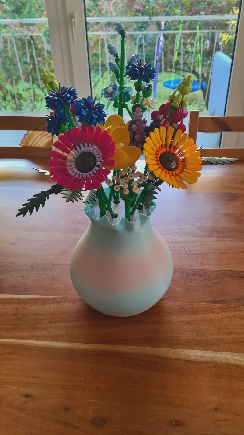 May include: A colorful bouquet of artificial flowers in a light blue and pink vase. The flowers include pink, yellow, and orange sunflowers, blue and purple blooms, and green stems. The vase sits on a wooden table.