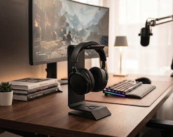 Puede incluir: Un soporte para auriculares en color negro mate con una base geométrica y un brazo de soporte vertical, que sostiene un par de auriculares negros de diadema. El soporte presenta un pequeño logotipo triangular en relieve.
