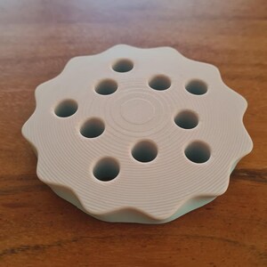 May include: A light gray, circular object with a wavy edge and ten evenly spaced holes. The object is made of a solid material and sits on a wooden surface. The design is simple and functional, possibly a tool or holder.