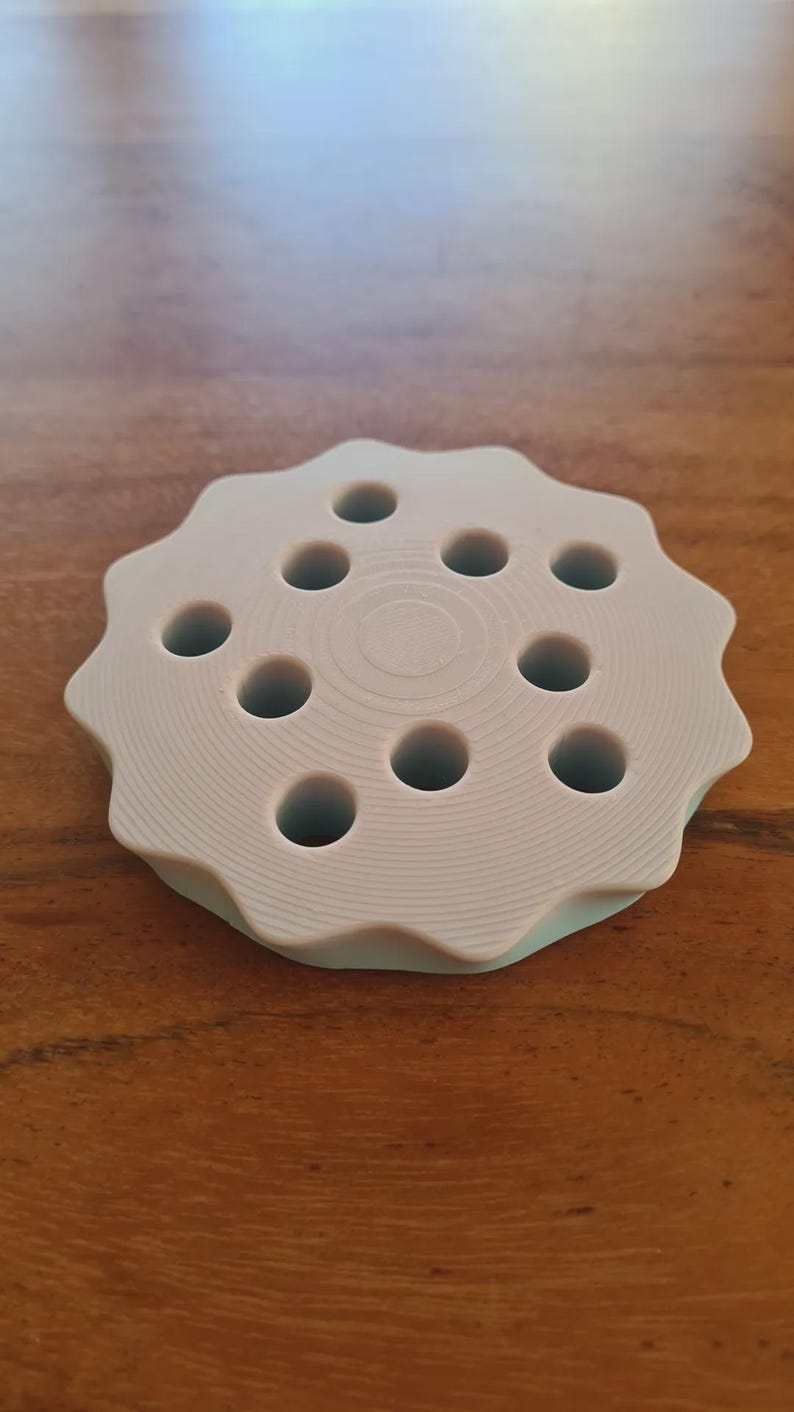 May include: A light gray, circular object with a wavy edge and ten evenly spaced holes. The object is made of a solid material and sits on a wooden surface. The design is simple and functional, possibly a tool or holder.