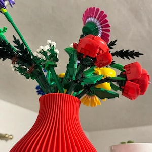 May include: A vibrant red vase holds a colorful arrangement of flowers. The bouquet includes red roses, yellow daisies, and purple irises, all with green stems and leaves. The vase has a ribbed design.