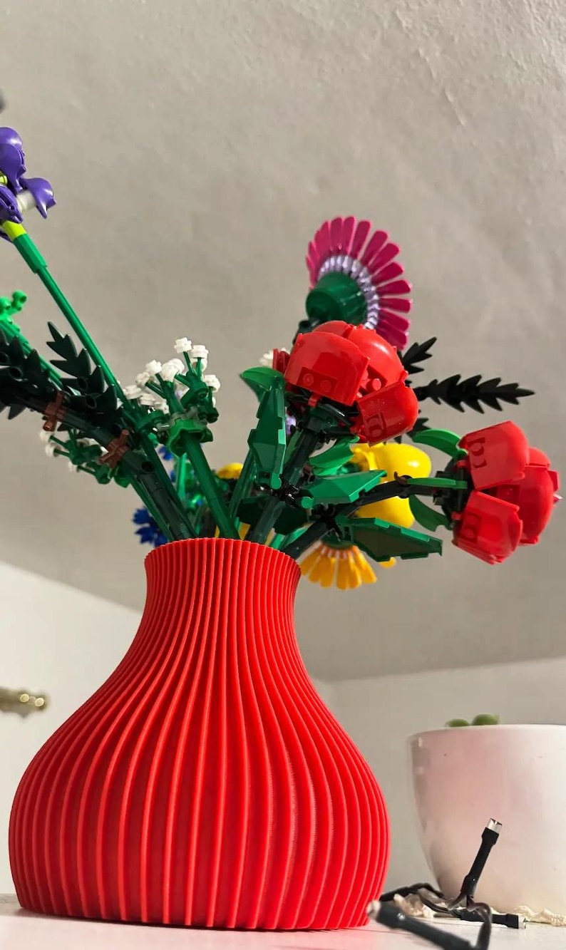 May include: A vibrant red vase holds a colorful arrangement of flowers. The bouquet includes red roses, yellow daisies, and purple irises, all with green stems and leaves. The vase has a ribbed design.