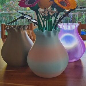 May include: Three decorative vases in shades of gray, blue, and purple, each with a ruffled rim. The vases hold a colorful arrangement of artificial flowers, including pink, yellow, and blue blooms. The vases are displayed on a wooden table.