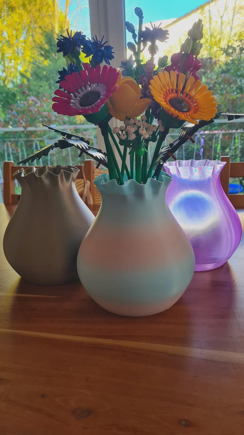 May include: Three decorative vases in shades of gray, blue, and purple, each with a ruffled rim. The vases hold a colorful arrangement of artificial flowers, including pink, yellow, and blue blooms. The vases are displayed on a wooden table.