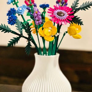 May include: A white, ribbed vase holds a vibrant bouquet of colorful flowers. The arrangement includes blue, pink, yellow, and purple blooms with green stems and leaves. The flowers are made of plastic and are arranged in a decorative display.