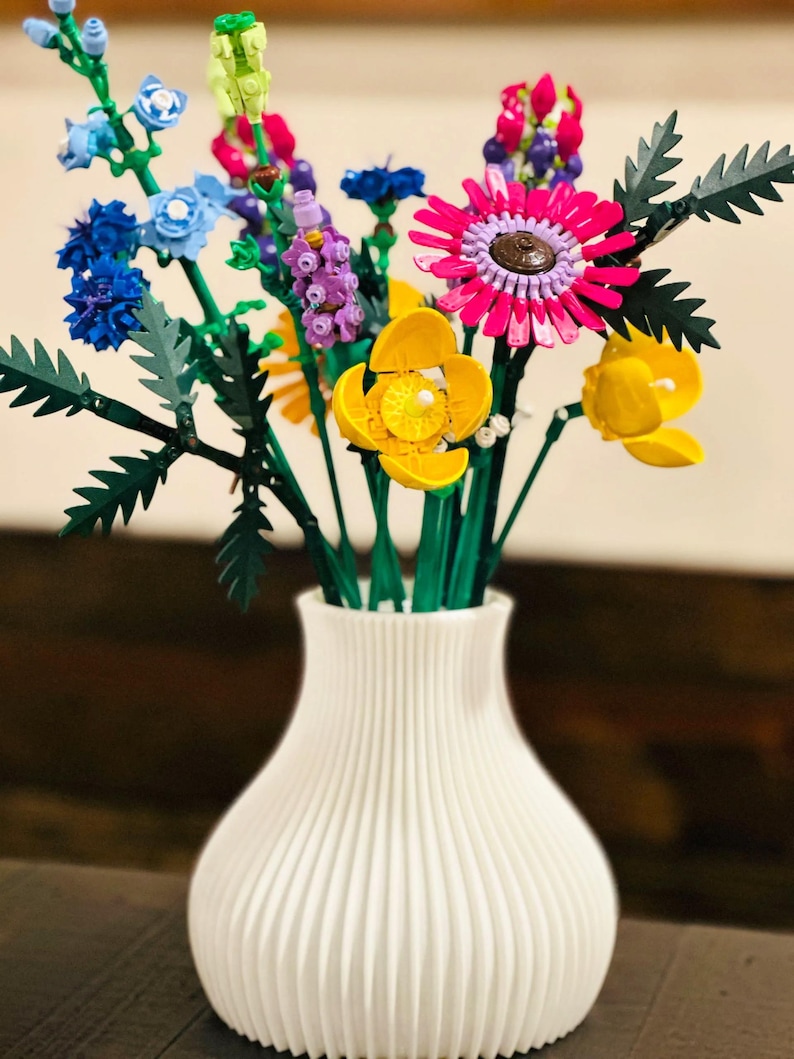 May include: A white, ribbed vase holds a vibrant bouquet of colorful flowers. The arrangement includes blue, pink, yellow, and purple blooms with green stems and leaves. The flowers are made of plastic and are arranged in a decorative display.