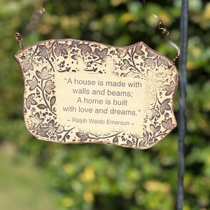 K&ouml;nnte beinhalten: Dekoratives Schild mit einem Zitat von Ralph Waldo Emerson, mit beigem Hintergrund und braunem Blumenrand. Auf dem Schild steht: &bdquo;Ein Haus wird mit Mauern und Balken gebaut; Ein Zuhause wird mit Liebe und Tr&auml;umen gebaut.&ldquo;