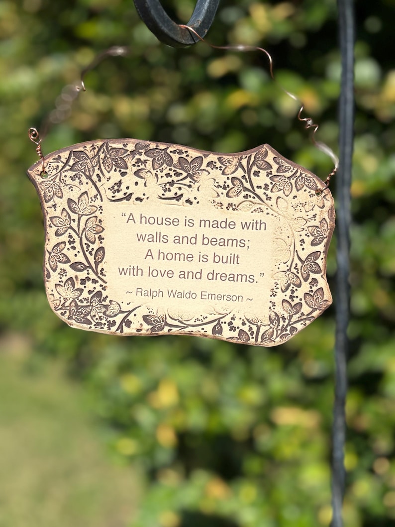 K&ouml;nnte beinhalten: Dekoratives Schild mit einem Zitat von Ralph Waldo Emerson, mit beigem Hintergrund und braunem Blumenrand. Auf dem Schild steht: &bdquo;Ein Haus wird mit Mauern und Balken gebaut; Ein Zuhause wird mit Liebe und Tr&auml;umen gebaut.&ldquo;