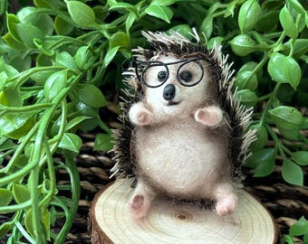 May include: A small, brown and white needle felted hedgehog wearing black framed glasses. The hedgehog is sitting on a small wooden slice.