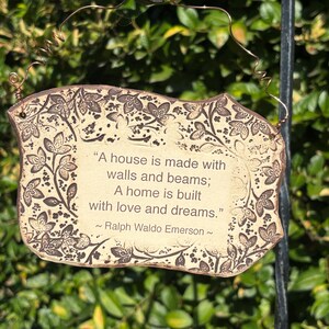 K&ouml;nnte beinhalten: Ein rechteckiges, cremefarbenes Schild mit braunem Blumenrand und Text. Auf dem Schild steht: &bdquo;Ein Haus wird mit Mauern und Balken gebaut; Ein Zuhause wird mit Liebe und Tr&auml;umen gebaut.&ldquo; Das Zitat wird Ralph Waldo Emerson zugeschrieben.