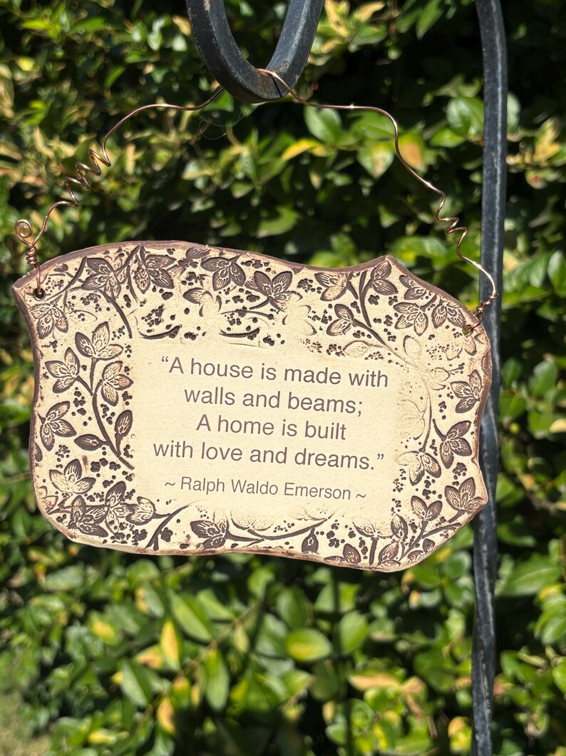 K&ouml;nnte beinhalten: Ein rechteckiges, cremefarbenes Schild mit braunem Blumenrand und Text. Auf dem Schild steht: &bdquo;Ein Haus wird mit Mauern und Balken gebaut; Ein Zuhause wird mit Liebe und Tr&auml;umen gebaut.&ldquo; Das Zitat wird Ralph Waldo Emerson zugeschrieben.