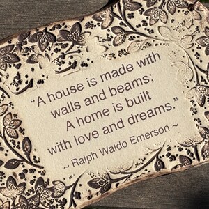 K&ouml;nnte beinhalten: Cremefarbene Keramikplakette mit floralem Rand und dem Zitat "A house is made with walls and beams; A home is built with love and dreams." von Ralph Waldo Emerson. Mit Kupferdraht zum Aufh&auml;ngen.