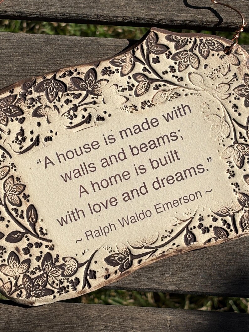 K&ouml;nnte beinhalten: Cremefarbene Keramikplakette mit floralem Rand und dem Zitat "A house is made with walls and beams; A home is built with love and dreams." von Ralph Waldo Emerson. Mit Kupferdraht zum Aufh&auml;ngen.