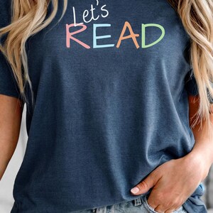 Puede incluir: Camiseta azul marino con el texto "Let's READ" en letras coloridas. La camiseta es de estilo cuello redondo y manga corta. La persona lleva pantalones cortos vaqueros.