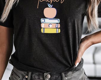 May include: Dark grey t-shirt featuring the word "Teacher" in cursive above a stack of books and an apple graphic. The books are blue, white, and yellow. The apple is peach-coloured.