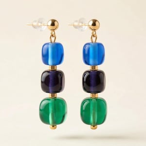 May include: A pair of drop earrings featuring three stacked, smooth, square-shaped glass beads in shades of blue, dark purple, and green. The beads are connected by gold-toned metal spacers and attached to a gold-toned post with a clear backing.