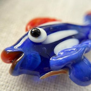 May include: A handcrafted glass fish figurine. The fish features a vibrant blue body with white stripes, a red fin, and a large, black-pupiled eye. The fish's mouth is a warm brown color.