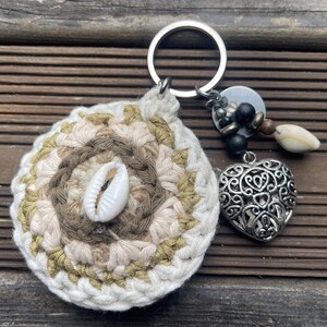 May include: A crocheted key chain with a white seashell, a silver heart charm, and a silver key ring. The crocheted circle is made with beige, brown, and green yarn.