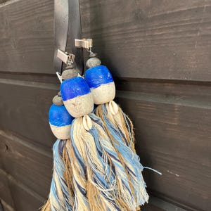 Può includere: Un fiocco blu e bianco a tema nautico con dettagli in legno e conchiglie. Il fiocco è appeso a un cinturino in pelle nera con clip argentate.