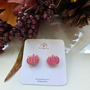 May include: A pair of pink pumpkin-shaped stud earrings with gold-colored detailing on a white display card. The card features the text The Sappy Pine and www.facebook.com/TheSappyPine. The earrings are presented against a background of autumn foliage.
